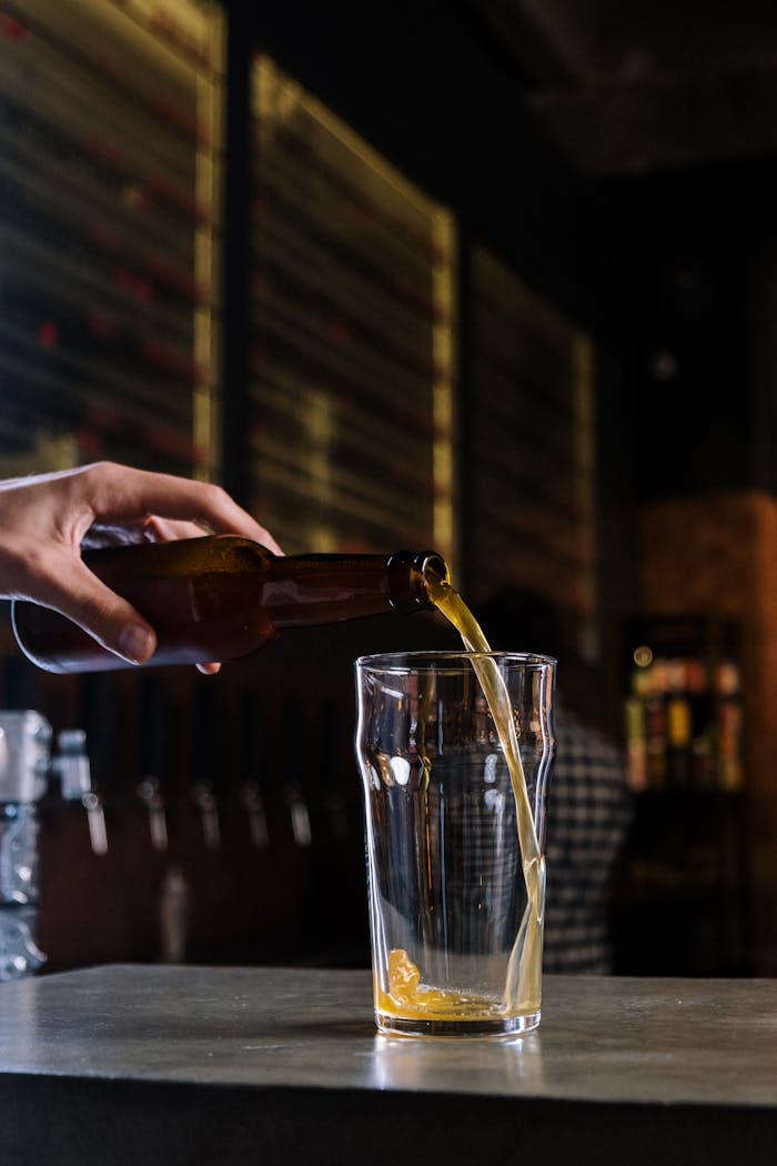 Hand pouring craft beer from a bottle into a glass in a dimly lit pub.